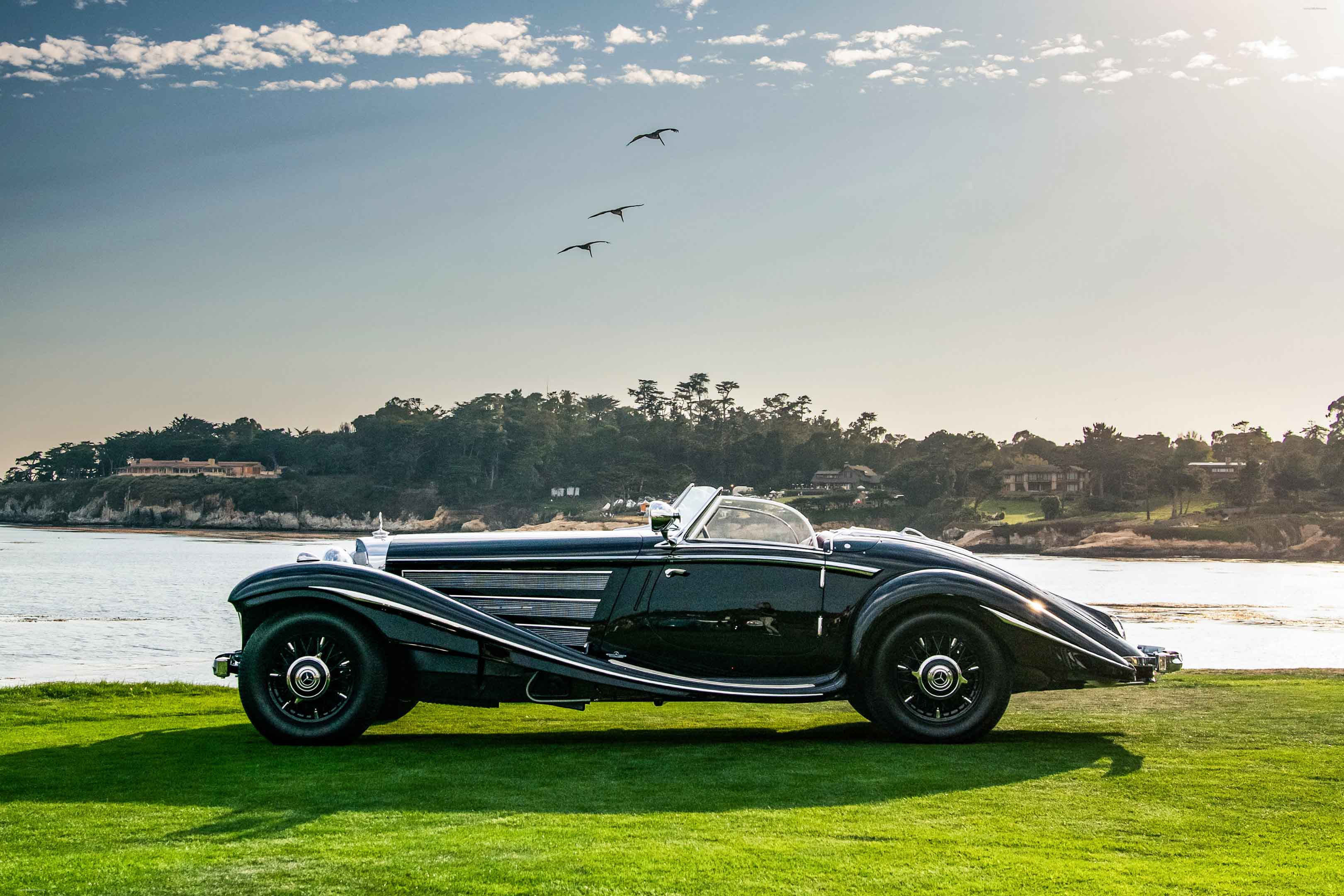 Black Mercedes Benz 540 K Special Roadster photographed beside the water, showing its long bonnet, sweeping fenders, chrome side trim and open cockpit.